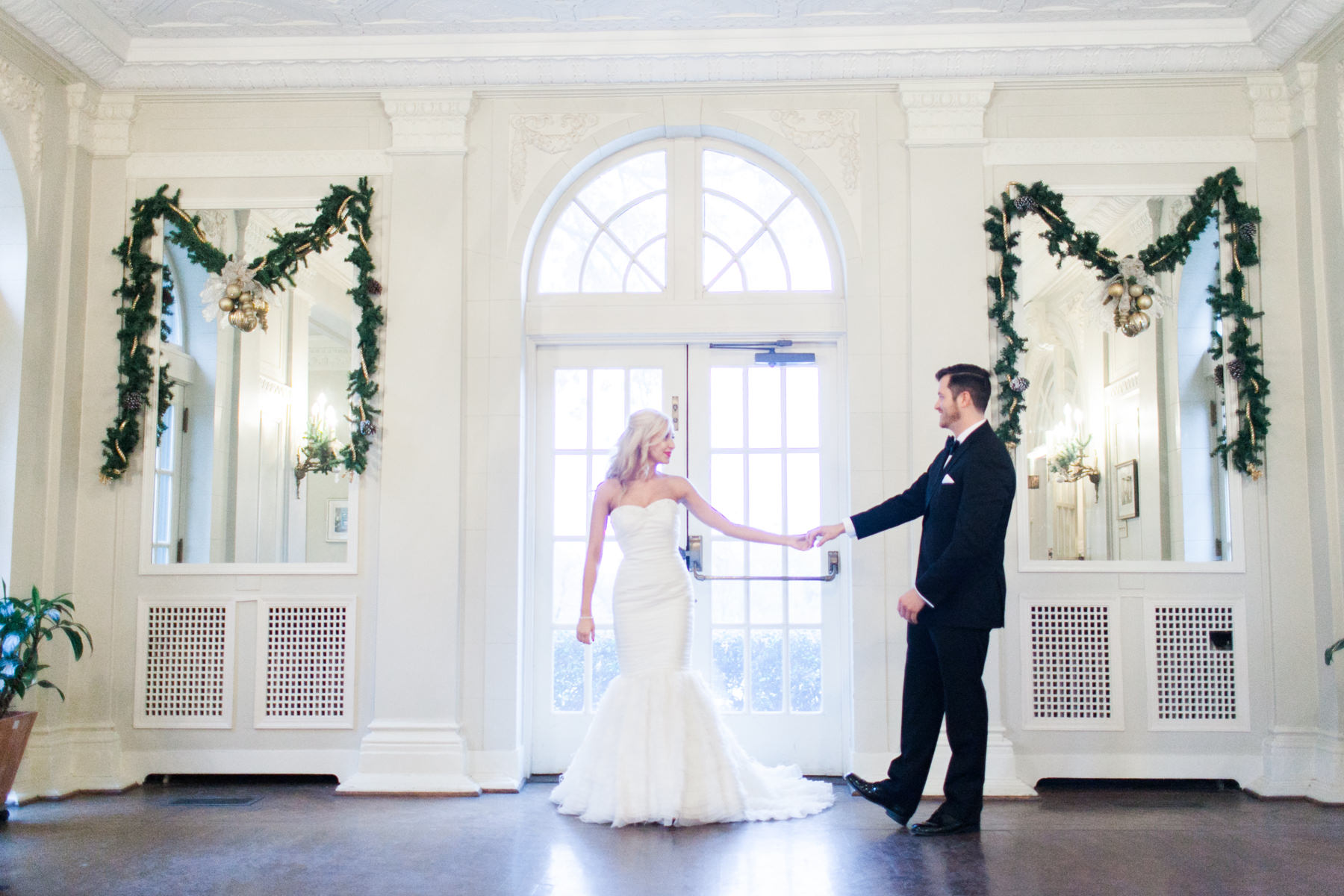 A bride and groom reaching for one another across a garlanded hall, the winter light coming straight through the doors behind them.