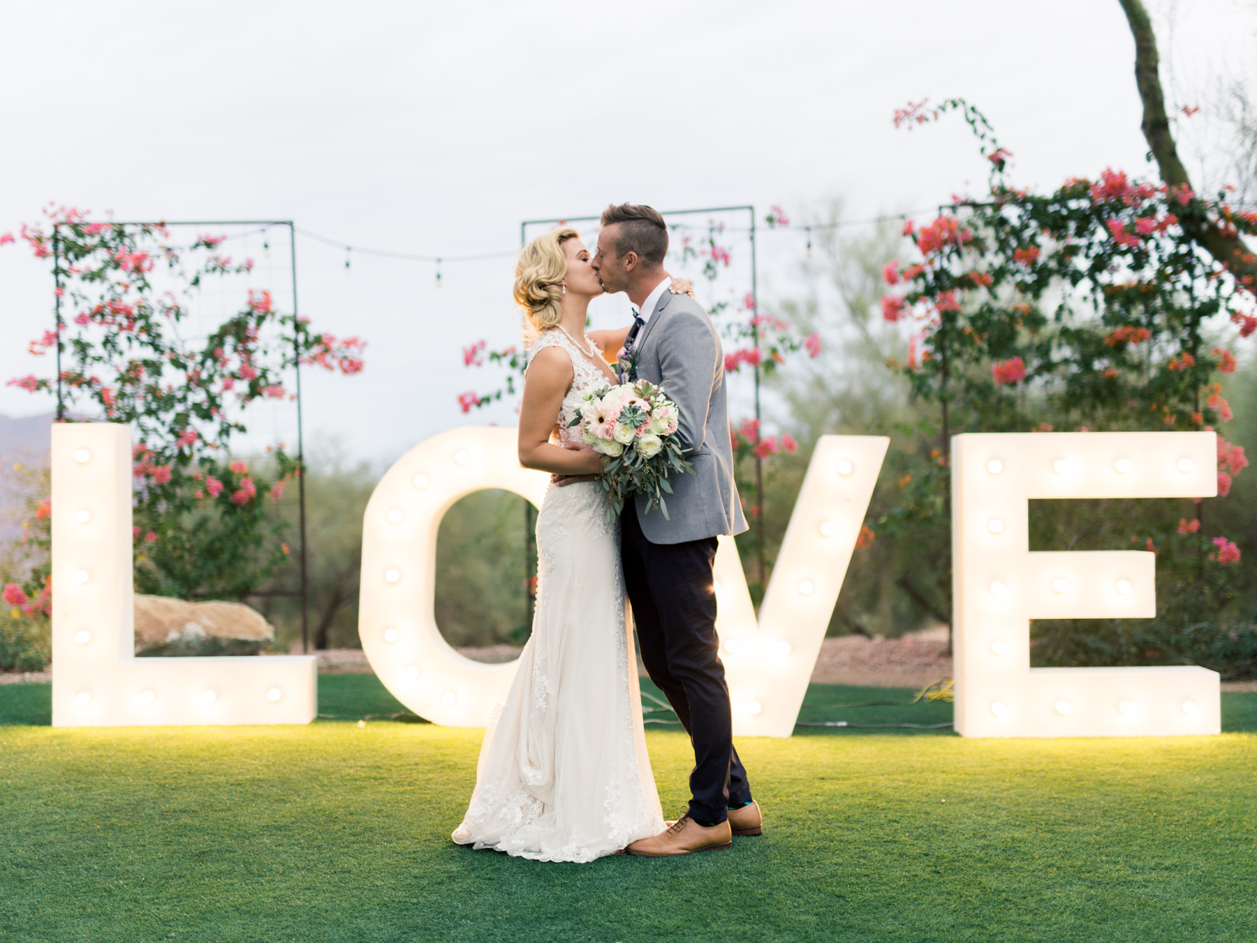 A kiss in front of lit marquee letters, with bougainvillea climbing the trellises behind.